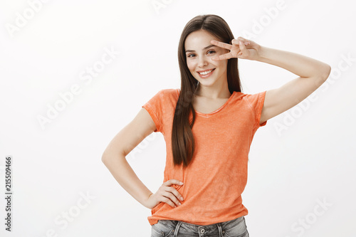 Everyone dance disco. Portrait of confident good-looking business woman in orange t-shirt, holding victory or peace sign over eye and smiling happily, standing over gray background