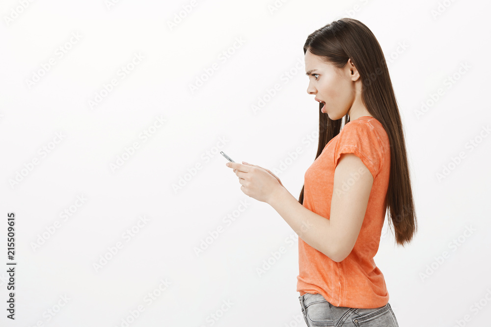 Profile portrait of shocked stunned expressive caucasian woman in orange t-shirt, holding smartphone and staring with confusion and revolt, seeing disturbing insulting message on screen over gray wall Profile portrait of shocked stunned expressive caucasian woman in orange t-shirt, holding smartphone and staring with confusion and revolt, seeing disturbing insulting message on screen over gray wall