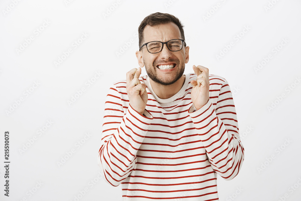 Portrait of nervous intense european man in black glasses, closing eyes and clenching teeth while holding fingers crossed and making wish, hoping or praying for fulfillment of dream Portrait of nervous intense european man in black glasses, closing eyes and clenching teeth while holding fingers crossed and making wish, hoping or praying for fulfillment of dream