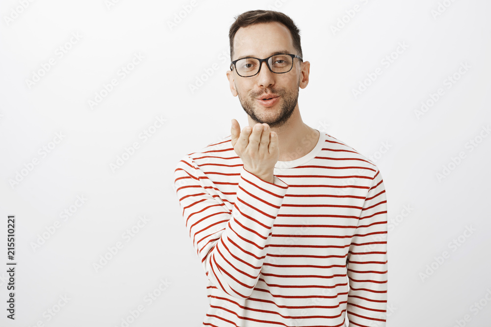 Sending kisses to you. Portrait of cute good-looking gay man in glasses,  holding palm near mouth and blowing mwah at camera with slight smile,  showing simpathy to boyfriend over gray wall Stock