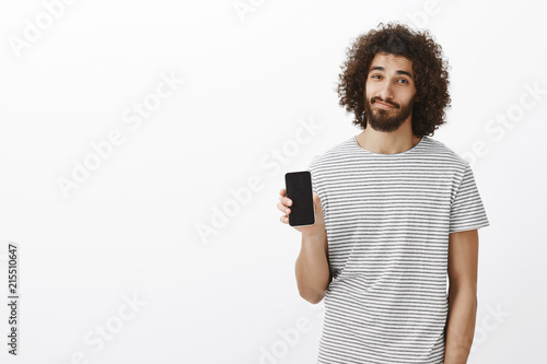 Indoor shot of calm casual attractive male coworker in striped t-shirt, showing black smartphone and smiling with relaxed pleased expression, offering nice device to cusomer over gray background photo