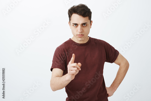 Indoor shot of disappointed strict adult brother, holding hand on hip and shaking index finger with threatening stare, looking from under forehead at camera, being angry and pissed off over gray wall © Cookie Studio