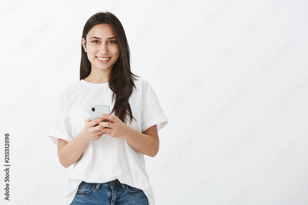 Portrait of friendly beautiful female student in white t-shirt, smiling broadly while holding smartphone and gazing at camera, listening carfully to teacher and making notes in gadget over gray wall Portrait of friendly beautiful female student in white t-shirt, smiling broadly while holding smartphone and gazing at camera, listening carfully to teacher and making notes in gadget over gray wall
