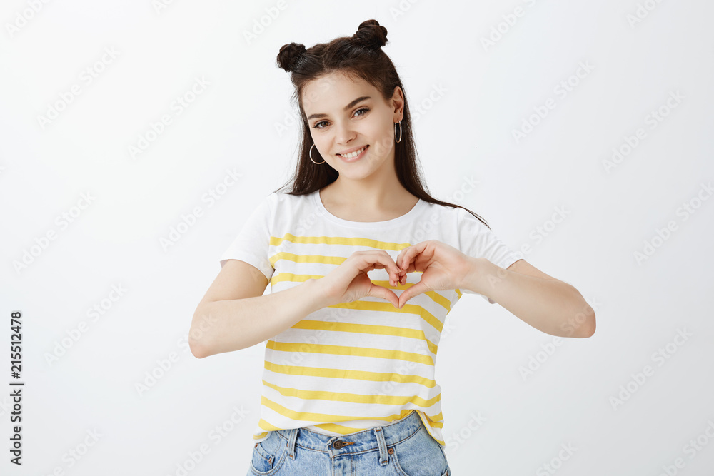 Feminine stylish caucasian girl in yellow striped t-shirt smiling joyfully and showing heart sign with hands over chest, expressing sympathy and like, standing sensual and flirty over grey wall Feminine stylish caucasian girl in yellow striped t-shirt smiling joyfully and showing heart sign with hands over chest, expressing sympathy and like, standing sensual and flirty over grey wall