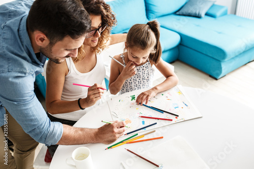 Parents  teach they young  daughter to draw.They sitting in living room and draws with colored pencils.
