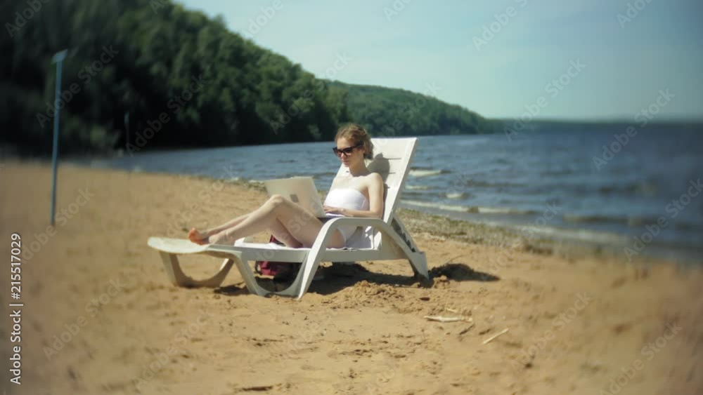 A young girl in a white bikini lies and tans on a deckchair on a sea sandy beach and is working on a laptop A young girl in a white bikini lies and tans on a deckchair on a sea sandy beach and is working on a laptop