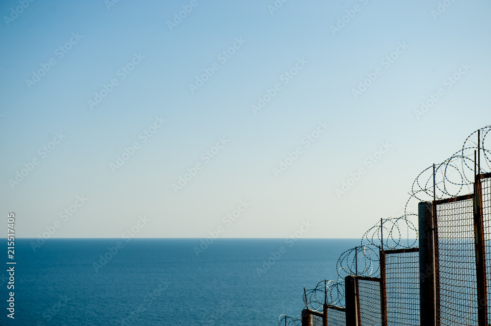 barbed razor fencing on state border against illegal immigration near sea sky - obrazy, fototapety, plakaty 