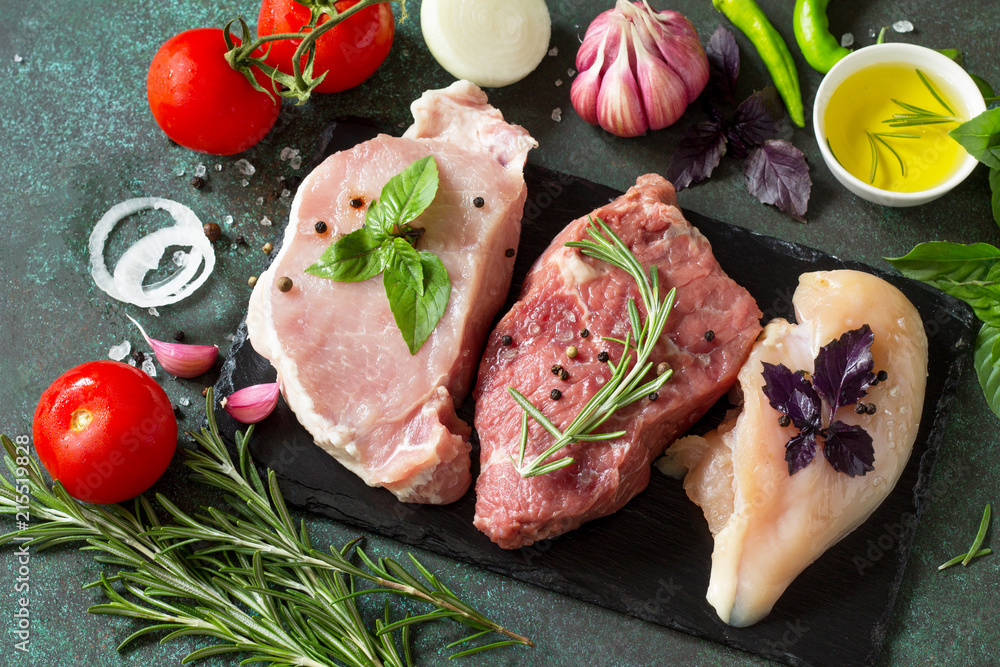 Fresh raw meat. Different types of raw pork meat, chicken fillet and beef with vegetables and herbs on dark wooden background.