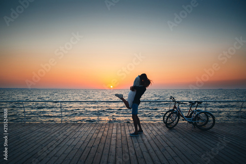 Man holding his girlfriend, couple of young lovers kissing at the beach at sunrise sky at wooden deck summer time © Iryna Budanova