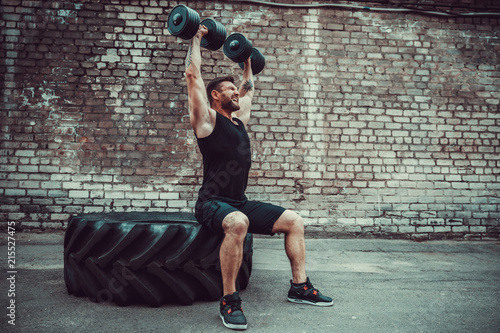 Athletic man working out with a dumbbell in front of brick wall. Strength and motivation. Outdoor workout. Exercise for the shoulder muscles, deltoid.