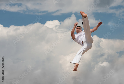 Young guy trains capoeira on sky backround. A men performs martial the kick in the jump