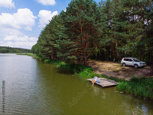 aerial view couple sitting at wooden doc looking at lake. suv on background