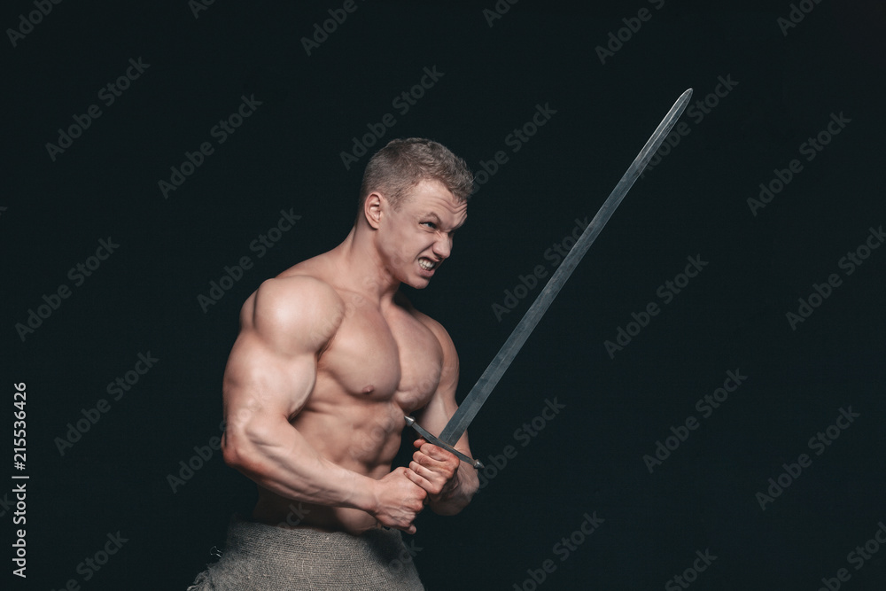 Bodybuilder man posing with a sword isolated on black background. Serious shirtless man demonstrating his mascular body Bodybuilder man posing with a sword isolated on black background. Serious shirtless man demonstrating his mascular body
