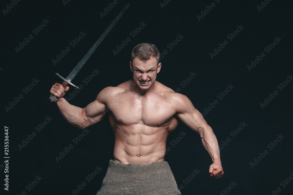 Bodybuilder man posing with a sword isolated on black background. Serious shirtless man demonstrating his mascular body Bodybuilder man posing with a sword isolated on black background. Serious shirtless man demonstrating his mascular body