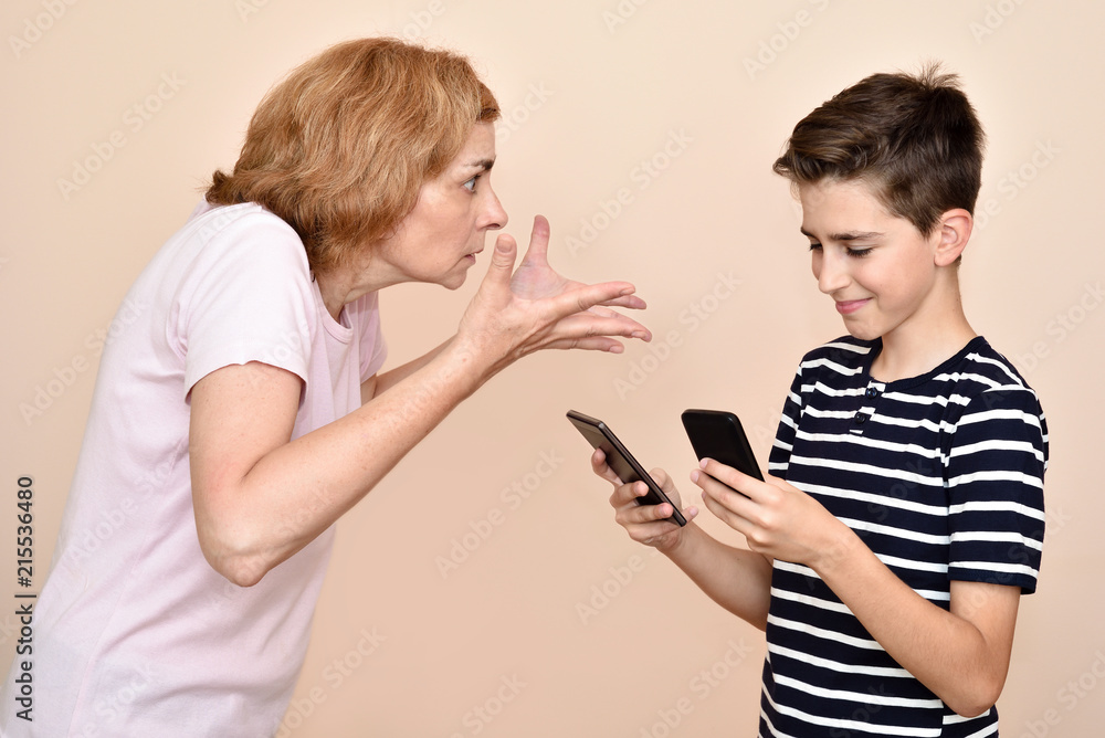Angry and nervous mother scolding her smiling son while he is looking at two smartphones at the same time. Family and modern technology addiction concept. Angry and nervous mother scolding her smiling son while he is looking at two smartphones at the same time. Family and modern technology addiction concept.