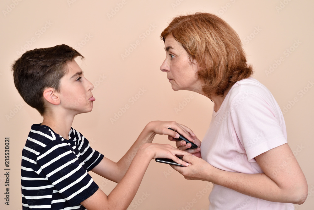 Pouting young boy does not allow his angry mother to check his two smarthphones. He is covering smarthphones with hands. They are looking each other. Family and modern technology addiction concept. Pouting young boy does not allow his angry mother to check his two smarthphones. He is covering smarthphones with hands. They are looking each other. Family and modern technology addiction concept.