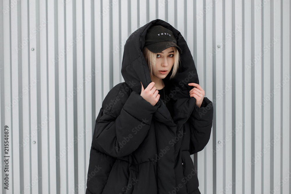 Fototapeta premium Fashionable beautiful model woman in black fashionable winter jacket and stylish cap posing near metal wall