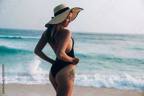 View from the back of a beautiful young girl on the beach pulling her bikini up, has slender legs and a rough sexy ass, wears a hat with wide brim and smiles.