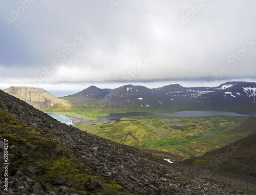 View on beautiful snow covered cliffs and fljotsvatn lake in Fljotavik cove in Hornstrandir, west fjords, Iceland, with river stream, green grass meadow, moody sky background photo