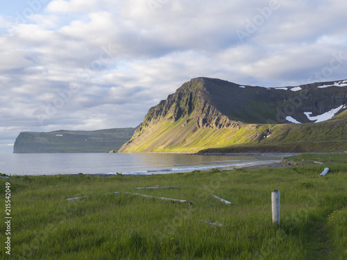 Hloduvik cove sea shore with green grass meadow, Skalarkambur mountain, wooden logs and footpath, blue sky white clouds background, Hornstrandir, west fjords, Iceland photo