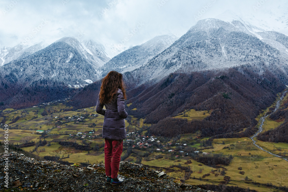 Young woman stands over a cliff and looks at snow-capped mountains, village in lower mount. Winter is coming, first snowfall, change of seasons. Georgia.