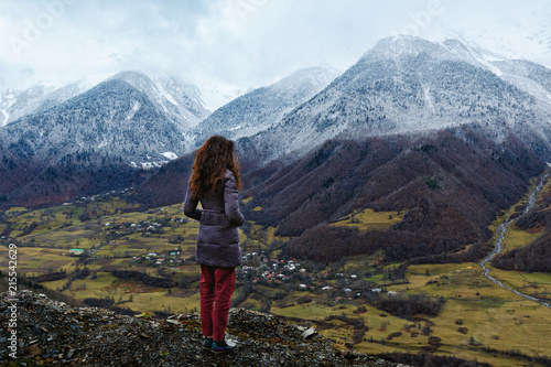 Young woman stands over a cliff and looks at snow-capped mountains, village in lower mount. Winter is coming, first snowfall, change of seasons. Georgia. © Lina