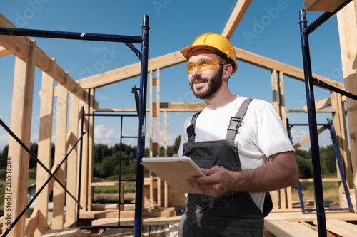 The man is a builder on the background of the roof of a frame house, in a yellow helmet and gray overalls. Uses a tablet photo