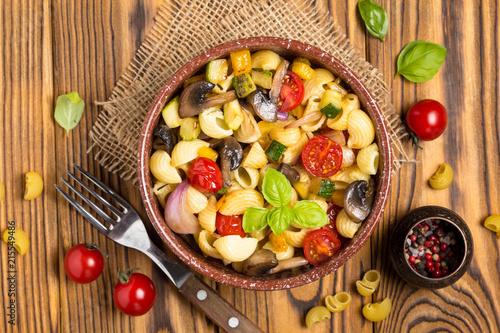 Italian pasta with fried vegetables and mushrooms, zucchini, cherry tomatoes, champignon, cheese and Basil. In dark bowl on wooden background