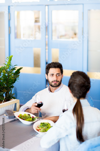 Young man with glass of red wine listening to his wife during talk by lunch