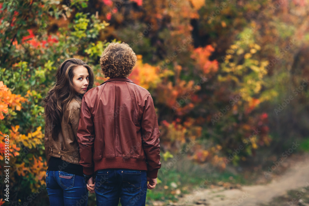 curly-haired man turned his back, and girl peeks out from behind his shoulder. loving couple on background of beautiful colorful autumn trees.