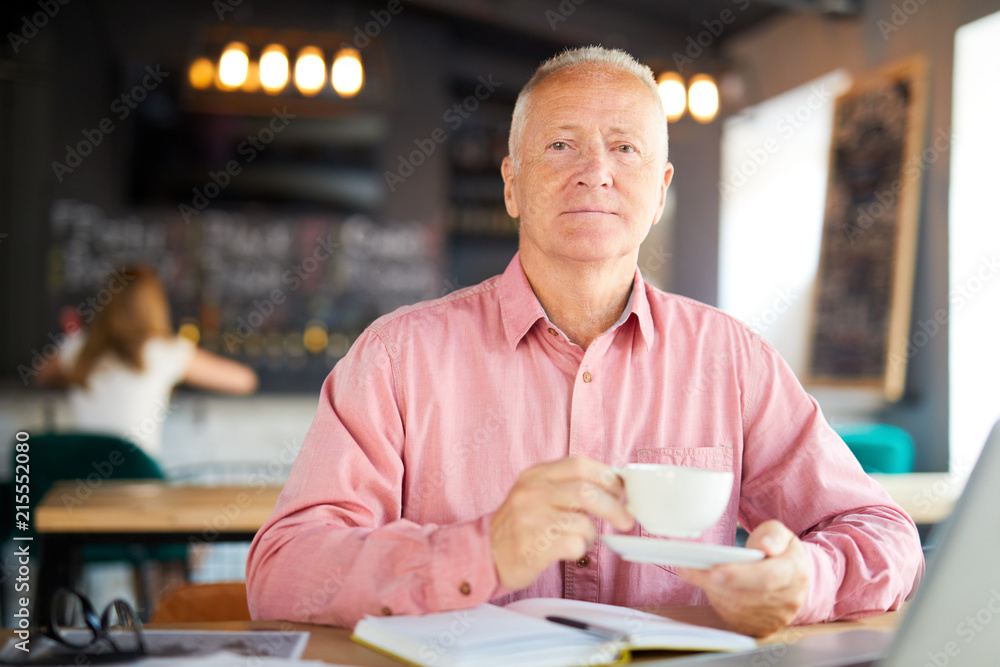 Serious senior man with cup of tea sitting by table in cafe while planning work at coffee-break Serious senior man with cup of tea sitting by table in cafe while planning work at coffee-break