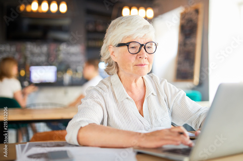 Senior grey-haired businesswoman sitting in cafe in front of laptop, typing and looking through online data