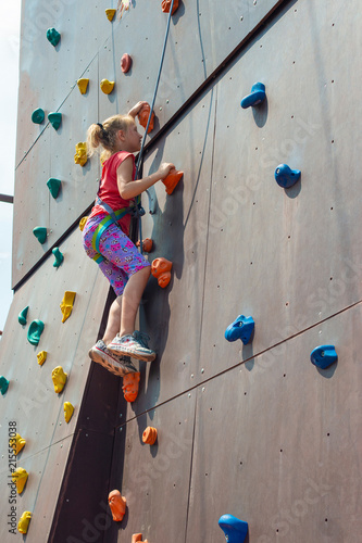 The girl is blonde, climbs to the top on an artificial rock with the help of a safety rope, is engaged in rock-climbing.