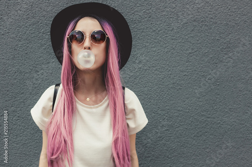 Stylish young hipster woman with long pink hair blowing a bubble with bubble gum.