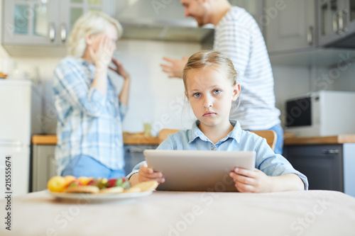 Unhappy little girl with touchpad looking at camera while her parents having argument