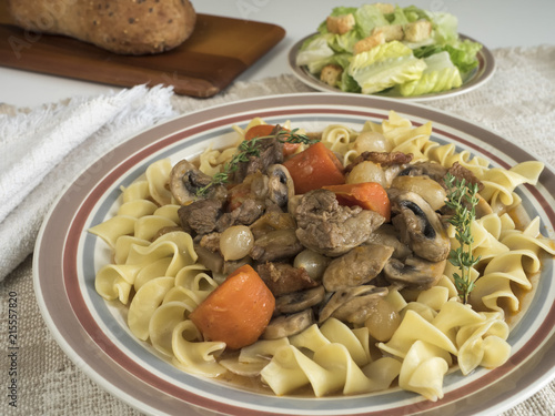 Plate of beef burgundy served over egg noodles with side of tossed green salad and baked bread