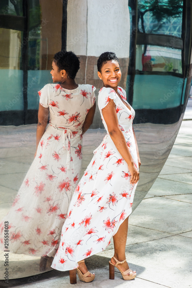 Young African American Woman with short afro hair, wearing long white patterned dress, standing by mirror in New York, turning around, smiling. Concept of self assured, self esteem, self checking..