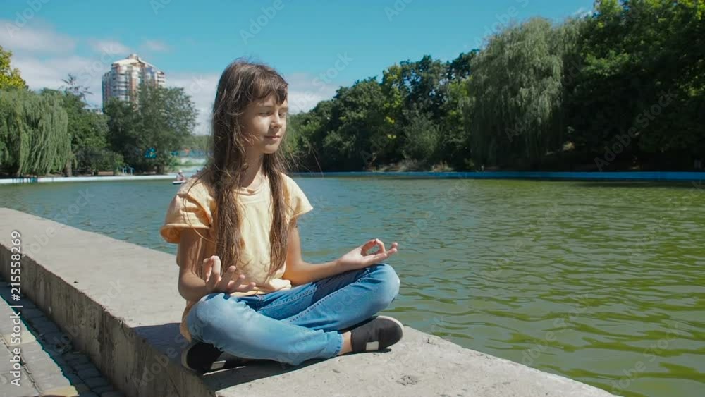 A child meditates in nature. A little girl in a lotus pose by the pond in the park. A child meditates in nature. A little girl in a lotus pose by the pond in the park.
