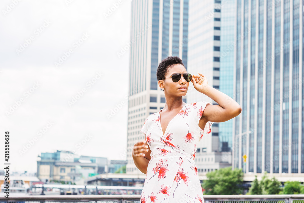 Young African American Woman traveling in New York, with short afro hair, wearing white patterned dress, sunglasses, standing in front of business district with high buildings of Manhattan, looking.. Young African American Woman traveling in New York, with short afro hair, wearing white patterned dress, sunglasses, standing in front of business district with high buildings of Manhattan, looking..