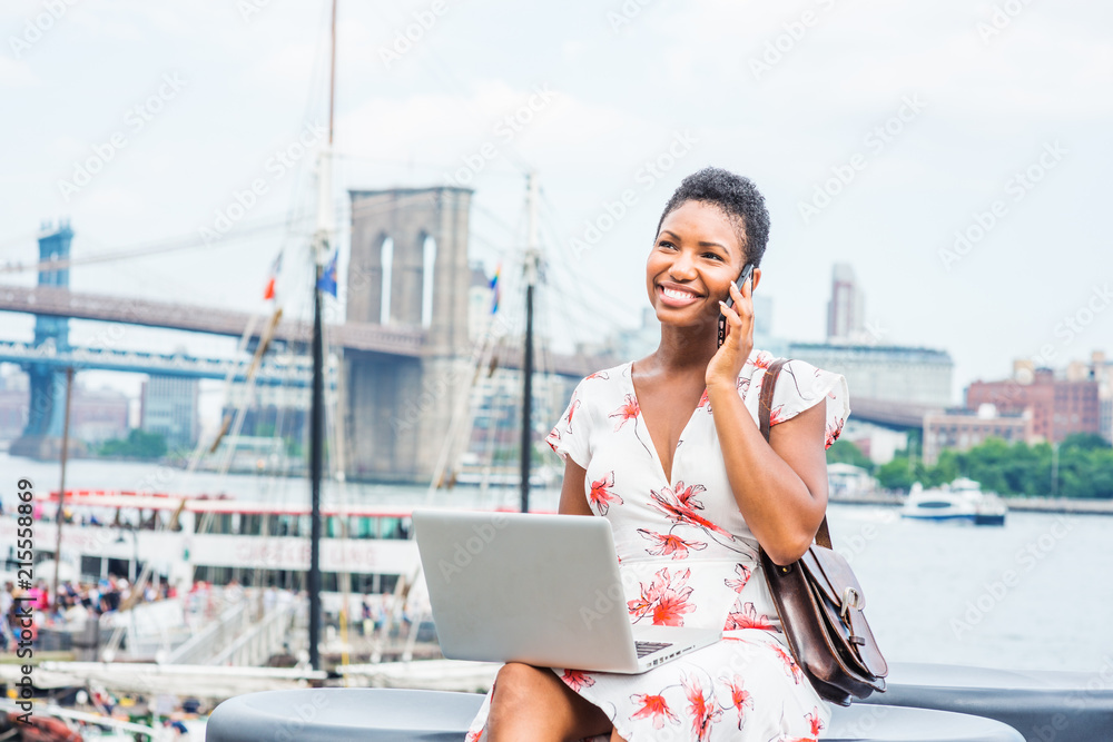 African American Woman with short afro hair traveling in New York, sitting by East River, talking on phone, working on laptop computer in same time. Manhattan, Brooklyn bridges, boat on background..