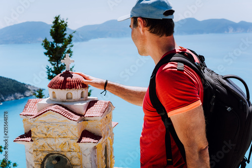 Male tourist touches thoughtful to small Hellenic shrine Proskinitari, Greece. Amazing sea view in the background photo