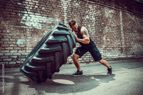 Muscular bearded tattooed fitness man moving large tire in street gym. Concept lifting, workout training.