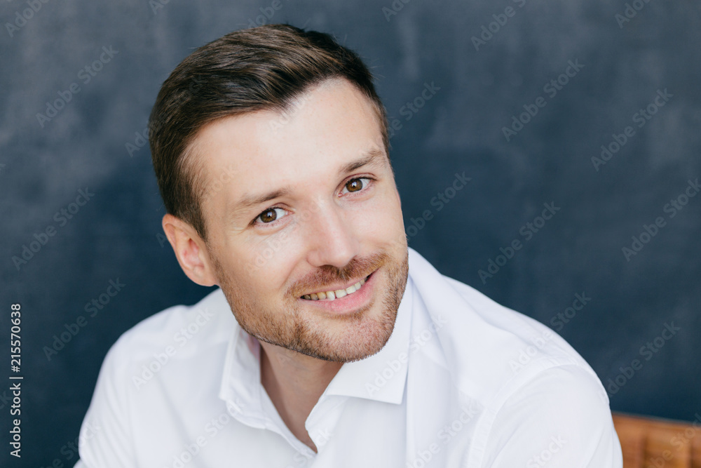 Photo of handsome unshaven male with bristle dressed in elegant white shirt, has charming smile, happy to meet with old friend, isolated over dark wall. People, emotions and lifestyle concept Photo of handsome unshaven male with bristle dressed in elegant white shirt, has charming smile, happy to meet with old friend, isolated over dark wall. People, emotions and lifestyle concept