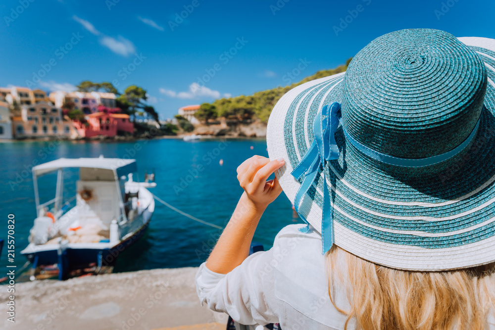 Happy young woman holding straw hat enjoying vacations in Assos village in front of emerald bay of Mediterranean sea white boat and beautiful traditional houses of in Kefalonia, Greece