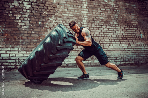 Muscular bearded tattooed fitness man moving large tire in street gym. Concept lifting, workout training. photo