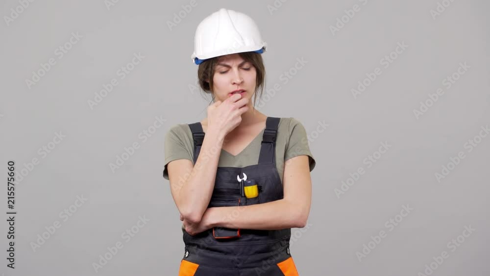 Portrait of brooding woman builder in helmet and jumpsuit touching chin and thinking while working on factory or manufacture, isolated over gray background Portrait of brooding woman builder in helmet and jumpsuit touching chin and thinking while working on factory or manufacture, isolated over gray background