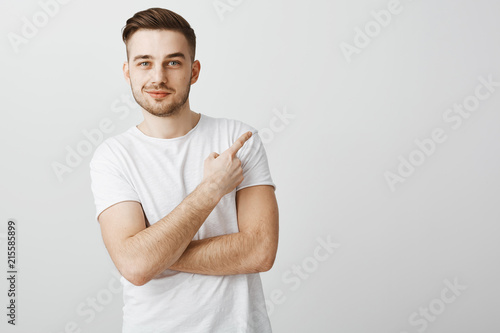 Studio shot of satisfied relaxed and handsome unshaven mature male with moustache pointing at upper right corner with delighted assured smirk standing over gray background