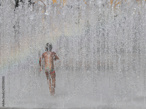 The boy joyfully runs and bathes in the city s fountain in the rays of the rainbow. Hot sunny weather. Children s carefree game in their spare time. Urban infrastructure. Refreshing splashes of water