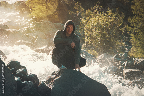 Male tourist sitting on a rock on the background of a waterfall surrounded by the splashing water. The guy is resting on the background of a mountain landscape with forest and fir trees photo