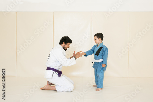 Father and little kid son are engaged in wrestling jiu-jitsu in the gym in a kimono. Trainer teaches child the methods and positions of single combat, karate or aikido.
